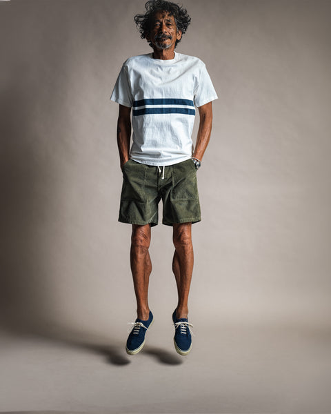 A man jumping wearing a Comp Stripe T-Shirt - Vintage White, olive green shorts, and navy sneakers.