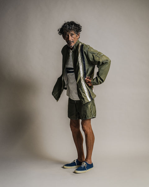 A man wearing a Racing Jacket 26 - Dark Olive/Bone/Black over a white t-shirt, paired with matching olive shorts and blue sneakers.