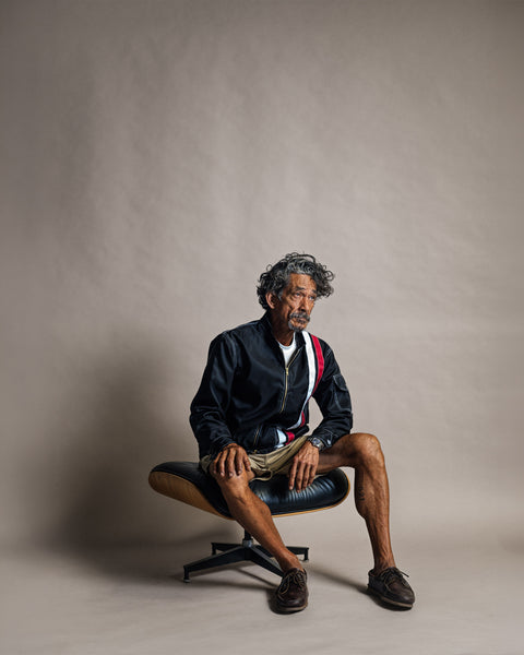 A man with grey curly hair sits on a lounge chair wearing a Racing Jacket 26 - Black/White/Red with red and white stripes on the shoulder, a white t-shirt, and tan shorts.