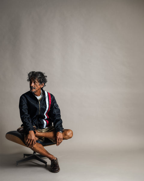 A man sits cross-legged on a chair wearing a Racing Jacket 26 - Black/White/Red, a vintage-inspired racing jacket with white and red stripes.