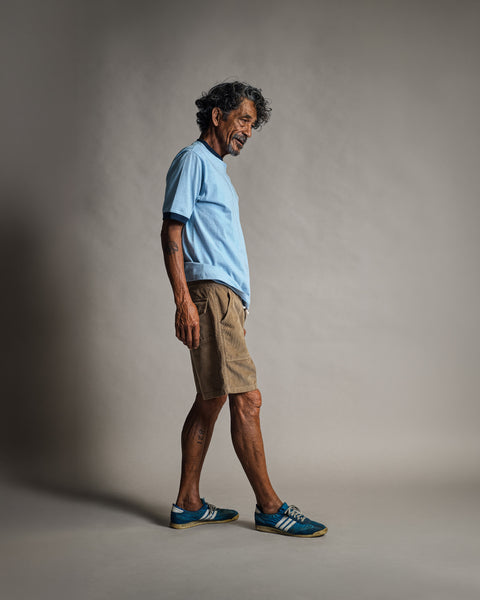 A man with dark curly hair and a mustache wearing a light blue t-shirt and Classic Corduroy Shorts - Sand, walking in profile against a grey background.