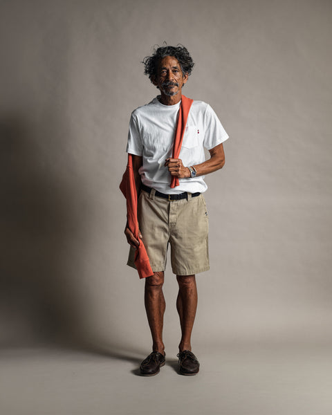 A man wearing a white t-shirt and Moss Stockman Chino Shorts stands holding a folded orange sweater over his shoulder.
