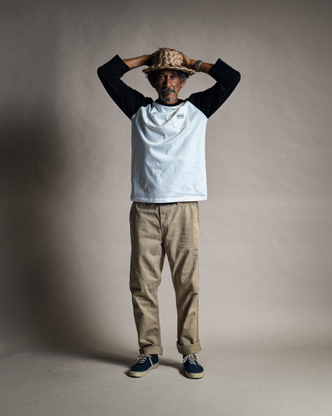 A man wearing a straw hat, a white and black baseball shirt, and Moss Stockman Chino Pants with the cuffs rolled.