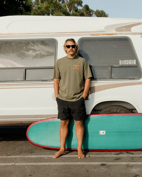 Kevin wearing an Army Green T-shirt with Birdwell License Plate Logo in orange over left of chest, standing in front of a teal surfboard with red trim and white vintage van parked in lot.