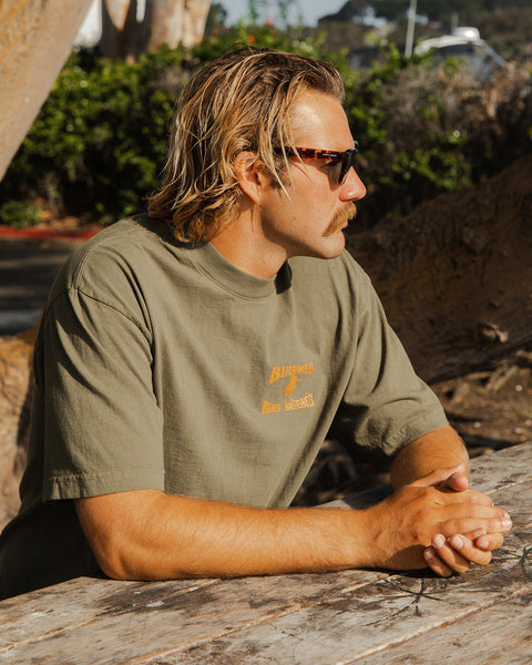 Man wearing sunglasses and a army t-shirt with orange license plate logo screen printed while sitting outdoors.