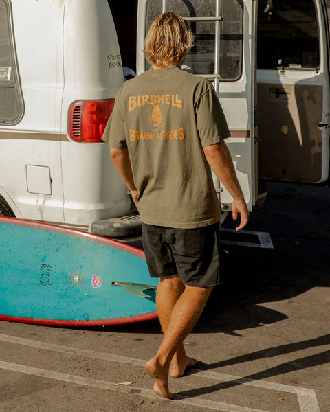 Kevin wearing an Army Green T-shirt with Birdwell License Plate Logo in orange, standing next to a blue surfboard and white vintage van parked in lot.