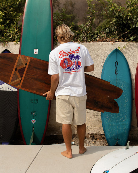 Person holding a surfboard with other boards stacked against a wall white wearing white Daydreamin Birdie T-Shirt.