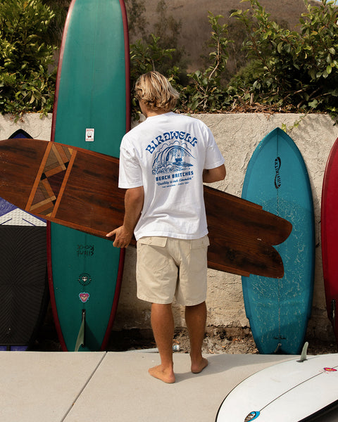 Person holding a wooden surfboard next to colorful surfboards against a wall while wearing a white t-shirt with blue Surfin Birdie graphic.