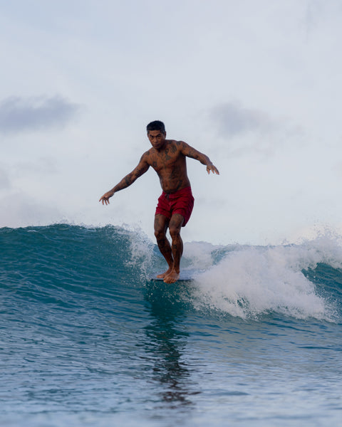 Mr. Toots glides on the nose of his surfboard in 310 Boardshorts in Red, arms out for balance, as a small wave breaks beneath him against a cloudy sky backdrop.