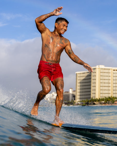 Mr. Toots, wearing 310 Boardshorts in Red, rides the nose of his surfboard with a big smile, balancing with one arm raised, as hotel buildings and a clear blue sky frame the background.