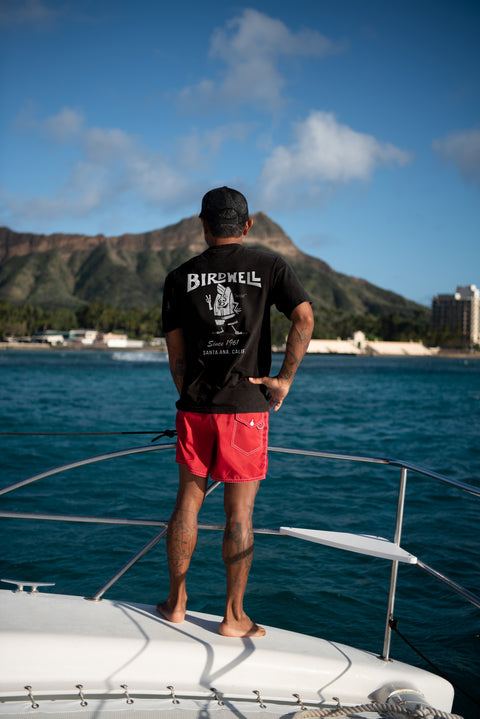 Toots wearing the 61 T-Shirt and 30 Boardshorts in Red while standing on the edge of a boat with mountain in the distance.