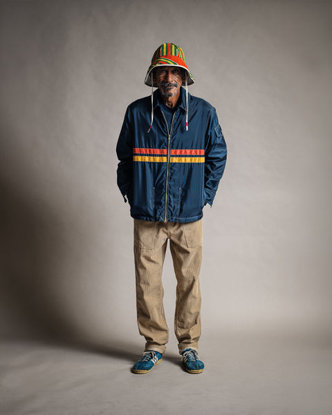 A man wears a Comp Jacket 26 - Navy/Gold/Paprika with horizontal yellow and orange stripes, tan trousers, and a multicolored bucket hat.