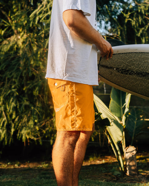 Side view of a person standing in a yard holding a surfboard, wearing the 300 Boardshorts in gold and a white t-shirt.