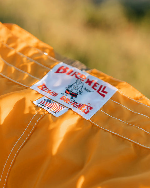 Side view of a person standing in a yard holding a surfboard, wearing the 300 Boardshorts in gold and a white t-shirt.