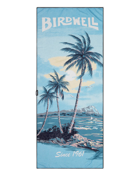The Postcard Beach Towel reads 'Birdwell Since 1961' and depicts a blue Hawaiian landscape illustration from Hoffman Fabrics.