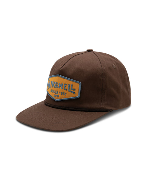 The Service Snapback in Brown, with embroidered gold and blue Birdwell patch and visor rope detail. Front angle.