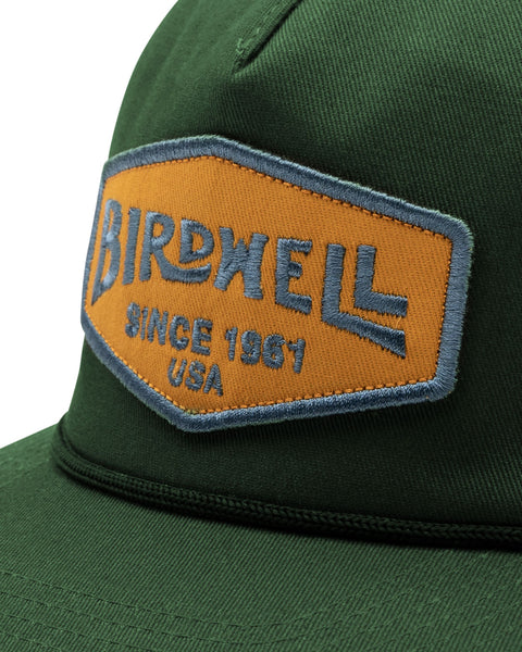 The Service Snapback in Green, with embroidered gold and blue Birdwell patch and visor rope detail. Close up image.