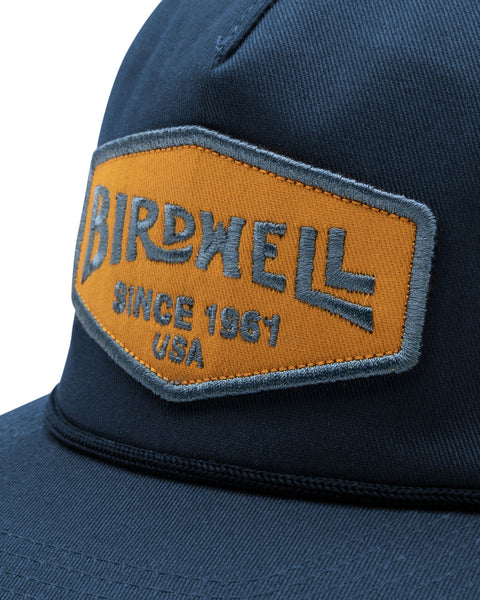 The Service Snapback in Navy, with embroidered gold and blue Birdwell patch and visor rope detail. Close up image.