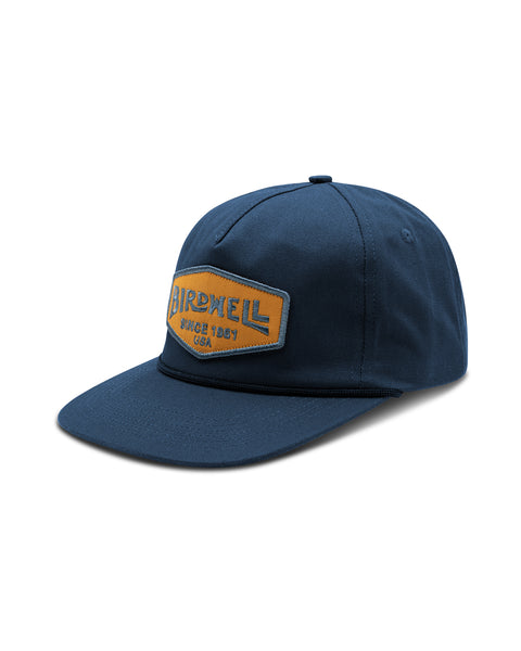 The Service Snapback in Navy, with embroidered gold and blue Birdwell patch and visor rope detail. Front angle.